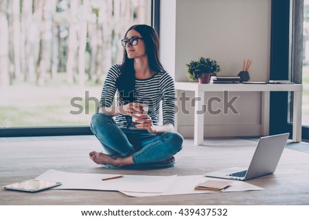 Just inspired. Confident young beautiful woman holding coffee cup and smiling while sitting on the floor at home with blueprint laying near her  Royalty-Free Stock Photo #439437532
