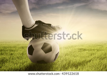 Picture of a soccer ball and foot of football player on the green meadow, shot at sunrise time