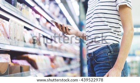 Young woman shopping healthy food in supermarket blur background. Close up view girl buy products using smartphone in store. Hipster at grocery using smartphone. Person comparing the price of produce