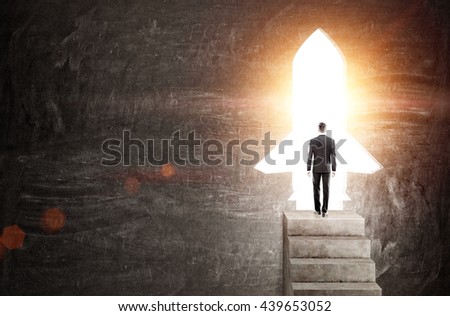 Startup concept with businessman standing on concrete stairs against rocket shaped door with bright light. Chalkboard background Royalty-Free Stock Photo #439653052