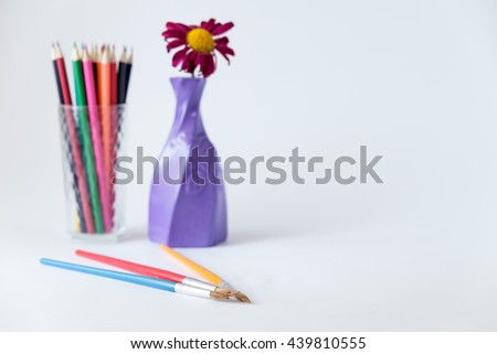 colorful pencils in glass cup near vase with flower and paintbrushes on foreground on white background 