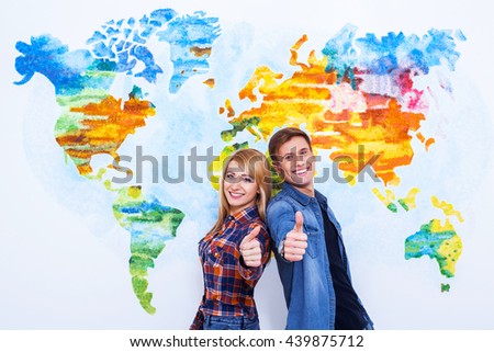 Everything will be great. Two happy young students showing their thumbs up and smiling while standing close to each other over wall painted like map of the world.