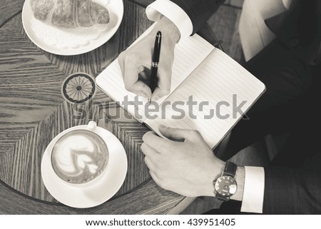 Close-up image of coffee break with a cap of coffee and croissant. Black and white, retro. Businessman writes something in notebook, office. Good Morning monday. Cafe, restaurant