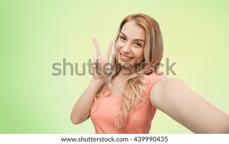 emotions, expressions and people concept - happy smiling young woman taking selfie and showing peace hand sign over green natural background