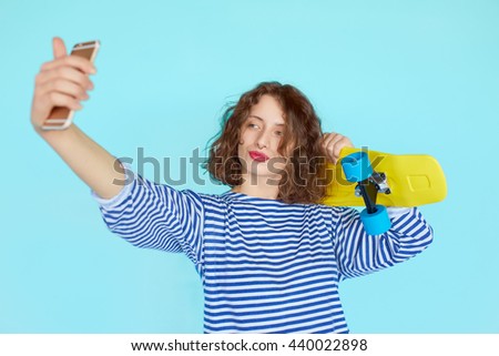 teenage girl in hipster outfit make selfie on a phone while holding a yellow skateboard isolated on bright colorful blue background
