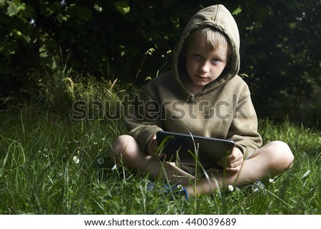 The blond boy is sitting on lawn and playing with the PC tablet