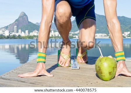 Track athlete with gold shoes crouching in starting blocks with coco gelado drinking coconut in front of the Rio de Janeiro skyline at Lagoa Rodrigo de Freitas lagoon