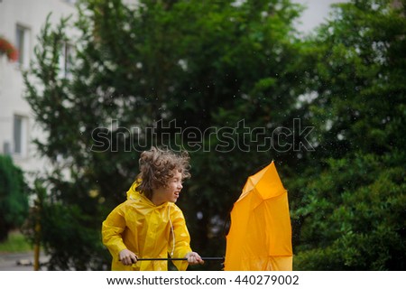 Laddie in a bright yellow raincoat and with an umbrella resists to rushes of strong wind. He hardly holds an umbrella. A fair hair of the boy was disheveled by wind. Royalty-Free Stock Photo #440279002