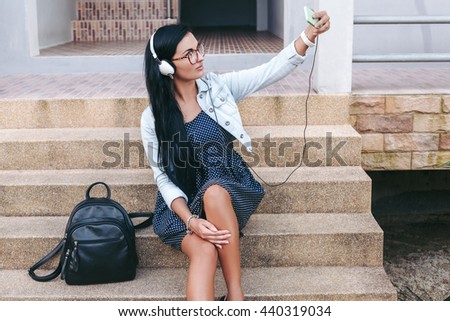 young stylish woman sitting on stairs with smartphone, listening to music on headphones, taking self photo