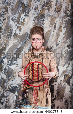 young girl with a pattern on the face and feathers looks straight into ethnic national style North American Indian background hairstyle and makeup holding a Dreamcatcher background