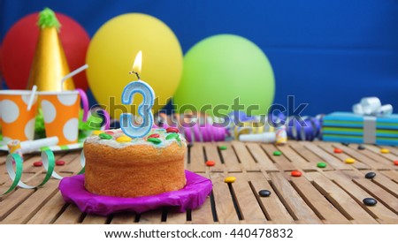 Birthday cake with candles on rustic wooden table with background of colorful balloons, gifts, plastic cups and candies with blue wall in the background. Focus is on cake