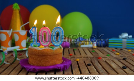 Birthday cake with candles on rustic wooden table with background of colorful balloons, gifts, plastic cups and candies with blue wall in the background. Focus is on cake