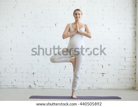 Pregnancy Yoga and Fitness concept. Portrait of beautiful young pregnant yoga model working out indoor. Pregnant happy fitness person practicing yoga at home. Prenatal Vrksasana Pose with Namaste