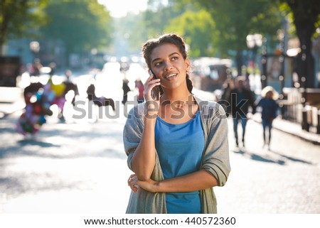 Cheerful charming african young girl dressed in blue t-shirt and grey jacket talked on her phone at the city background.