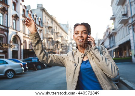 Attractive girl dressed in blue t-shirt and grey jacket talking on the phone, hold the book and catching the car at the street background.