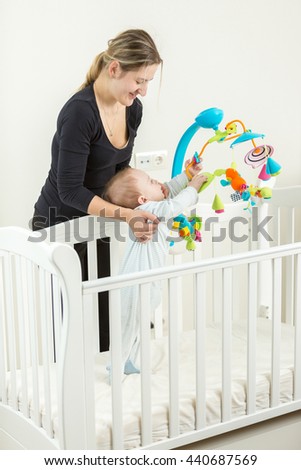 Happy young mother holding cute baby boy standing in white wooden cot