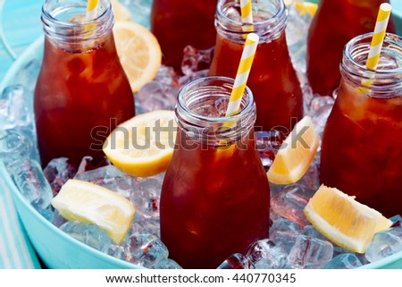 Close up of glass milk bottles filled with iced tea and fresh lemon with yellow swirled straw on ice in round blue metal tub sitting on bright blue wooden table with blue and white striped napkin