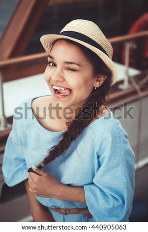 Closeup portrait of funny beautiful smiling white Caucasian brunette girl winking showing tongue, in blue dress and straw hat, by yacht boat, navy maritime retro vintage style concept