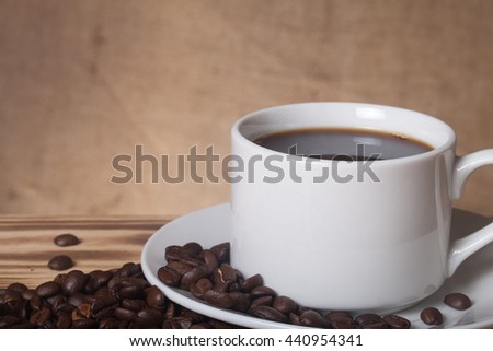 Coffee beans and coffee in white cup on wooden table opposite a defocused burlap background.
