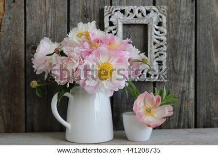 Composition with softness pastel bouquet of pink peonies in white ceramic pitcher, small cup , flower pot with one peony, vintage wooden white rectangular frame on the dark wooden background. 