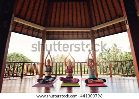 Three young women practicing yoga in a gym sitting cross legged on their mats and meditating. Group of people doing meditation in yoga class.