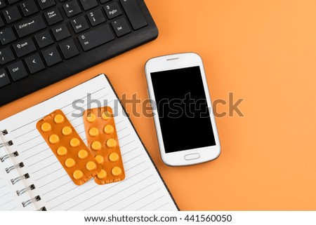 Office desk table with white smart phone, black computer keyboard, blank spiral notebook and orange blisters of pharmaceutical pills on orange desk background