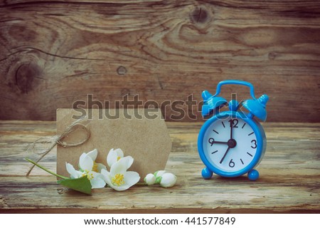 
Blue alarm clock, empty paper tag, jasmine branch, old wooden table, retro style