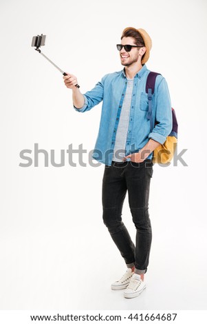 Full length of smiling young man with backpack taking photos with smartphone and selfie stick