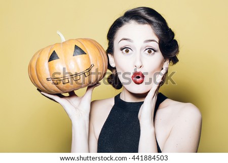 Surprised woman with beautiful face and retro hairdo and red lips holding pumpkin in studio on yellow background. Halloween concept Royalty-Free Stock Photo #441884503