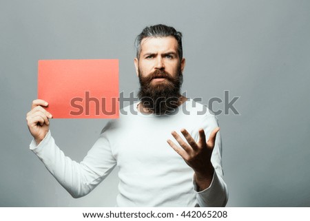 young bearded man with serious face holding red paper sheet in studio on grey background, copy space