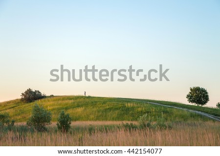 Red sunset over green hill with road. Road lane and cloudless sky. Nature design. Rural landscape in countryside. Rolling hill, trees, bushes, green field on warm sunset. Path turns uphill to the sky.