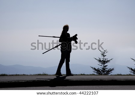 Silhouette of woman photographer carrying a tripod and camera while looking out over a mountain vista.