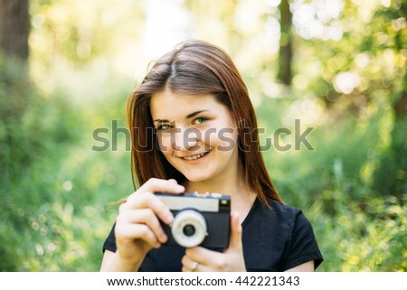 Happy Red-haired Caucasian Girl Young Woman Photographer Taking Pictures The Old Retro Vintage Film Camera In Summer Green Forest. Girl Dressed In A Black T-shirt