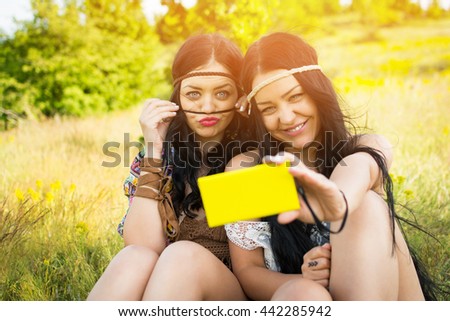 Two cute cheerful sisters in boho hippie outfits taking a selfie on smartphone, outdoors on sunny summer day. Closeup of young women making funny faces, photographing themselves using cellphone.