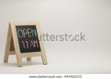 A hand writing 'Open 11 AM' on chalkboard. small wooden vintage style blackboard on white gradient background. colorful chalk.Text space on right screen for web template or presentation or marketing.