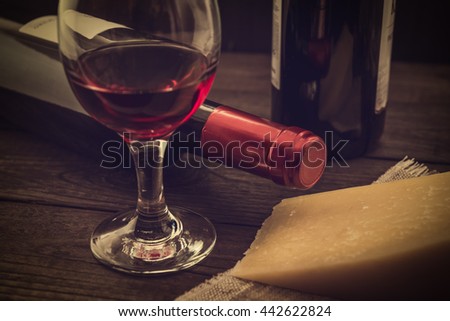 Two bottles of red wine with a glass of red wine and a piece of parmesan on an old wooden table. Angle view, focus on the bottle of red wine, image vignetting and the yellow toning