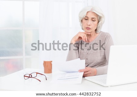 Photo of beautiful emotional adult woman. White interior with window. Upset woman working with bills