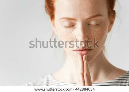 Beautiful Caucasian redhead woman praying or practising yoga at home, holding hands in namaste. Student girl with healthy freckled skin, closing her eyes, holding hands in prayer, hoping for the best Royalty-Free Stock Photo #442884034