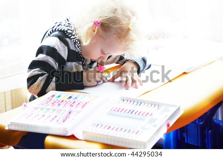 Little girl draws a colored pencil sitting at a table looking at the child a sheet of paper