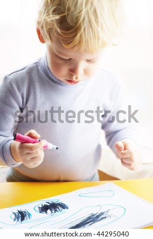 Little boy draws a colored pencil sitting at a table looking at the child a sheet of paper