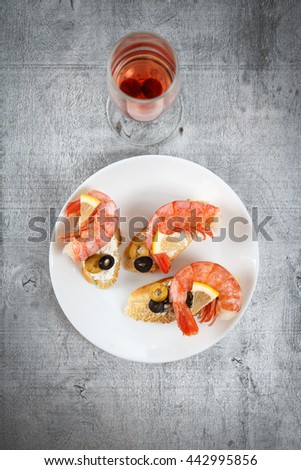 Tasty various italian sandwiches with seafood against rustic wooden background. Crostini with cheese, king shrimps, lemon, sliced olives on white plate and glass of wine, top view