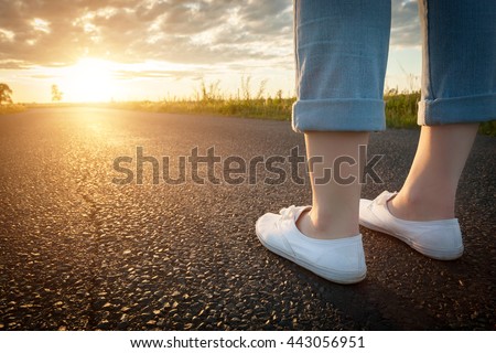 Woman in white sneakers standing on asphalt road towards sun. Concept of new start, travel, freedom etc. Royalty-Free Stock Photo #443056951