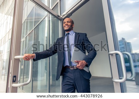 Low angle view of handsome young Afro American businessman in classic suit holding a laptop and smiling while leaving the office building Royalty-Free Stock Photo #443078926
