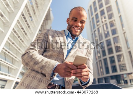 Handsome young Afro American businessman in glasses is using smartphone and smiling, standing outdoors