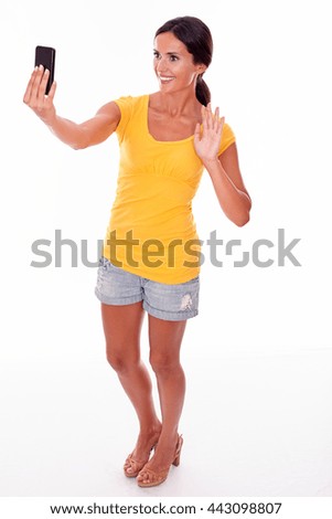 Smiling brunette holding a cell phone looking at the phone for a selfie while wearing a yellow t-shirt and short jeans isolated