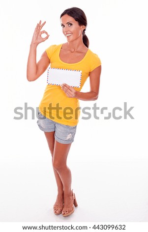 Smiling brunette woman gesturing a perfect sign holding a blank envelope while looking at camera wearing a yellow t-shirt and short jeans isolated