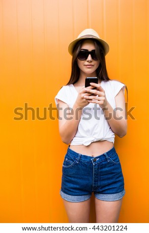 Pretty woman in sunglasses and hat text message on phone over orange colorful wall background
