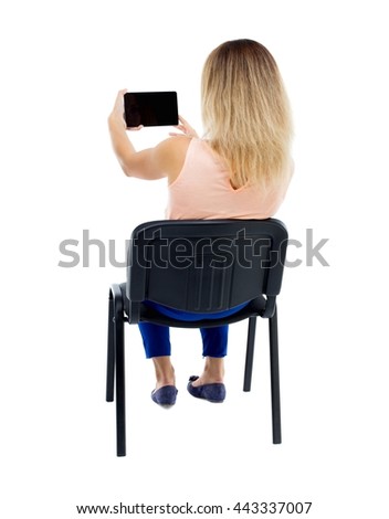 back view woman sitting on chair and looks at screen of the tablet.  Rear view people collection  backside view person. Blonde in blue trousers sitting on office chair and shoots video smartphone
