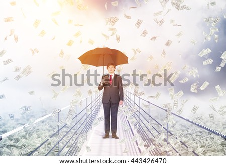 Financial growth and success concept with businessman holding umbrella and standing in the middle of bridge with abstract dollar banknote rain on cloudy background with sunlight