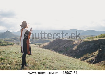 Young beautiful woman traveler wearing hat and poncho taking pictures on her smart phone standing on the top of the mountain